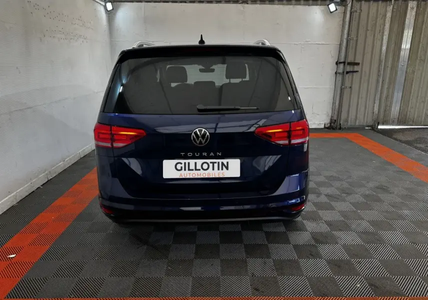 Vue arrière d'un Volkswagen Touran bleu 2024 garé dans un garage avec sol à damier noir et orange.