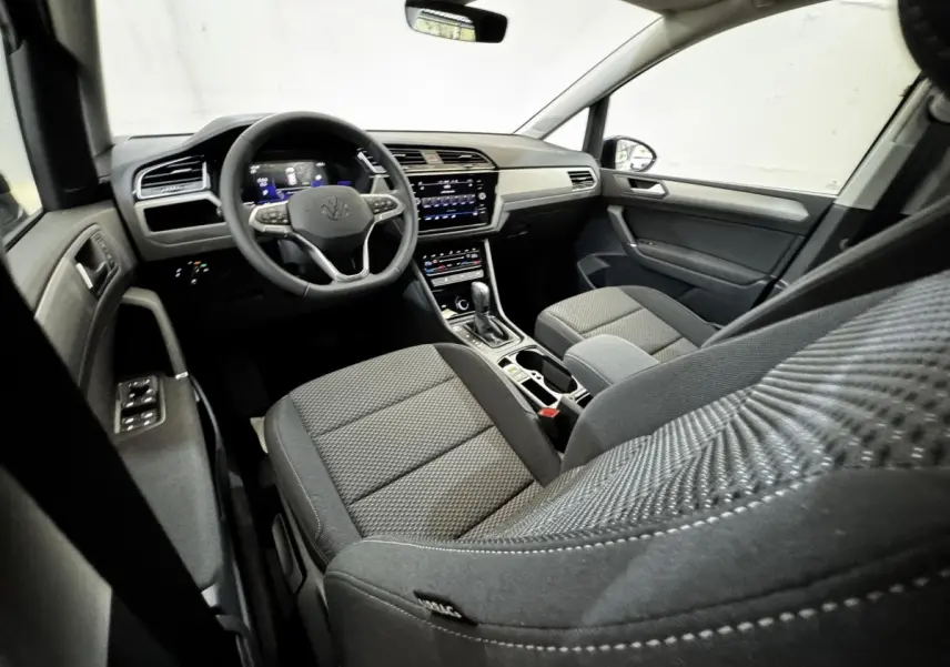 Vue intérieure côté conducteur du Volkswagen Touran 2024, montrant tableau de bord et sièges tissu gris foncé.
