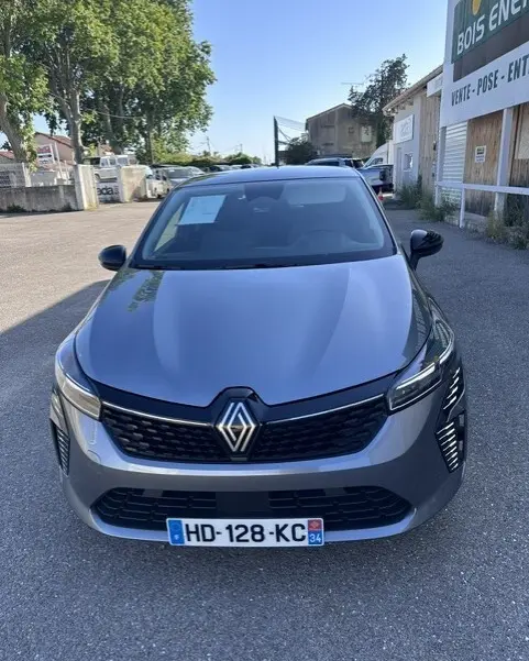 Vue avant d'une Renault Clio gris métallisé 2025, avec calandre noire et phares LED allumés, stationnée en extérieur.