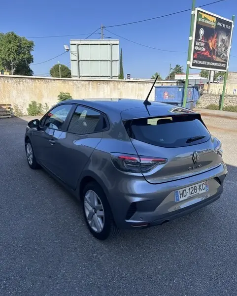 Renault Clio gris 2025 vue 3/4 arrière droit, vitres surteintées et antenne sur le toit dans un parking extérieur.