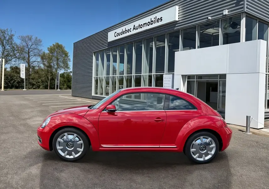 Profil droit d'une Volkswagen Coccinelle rouge Tornade 2012, avec jantes 18 pouces et carrosserie brillante devant un concessionnaire.