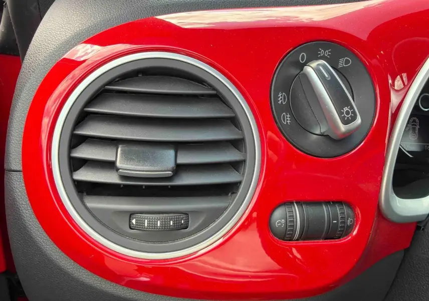 Détail du tableau de bord rouge brillant côté gauche avec commande des phares et bouche d’aération ronde sur Volkswagen Coccinelle 2012.