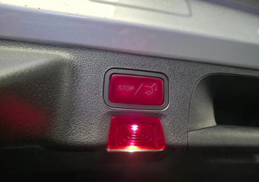 Gros plan sur le bouton rouge d'ouverture/fermeture automatique du hayon dans le coffre d'une Mercedes CLA Shooting Brake gris.
