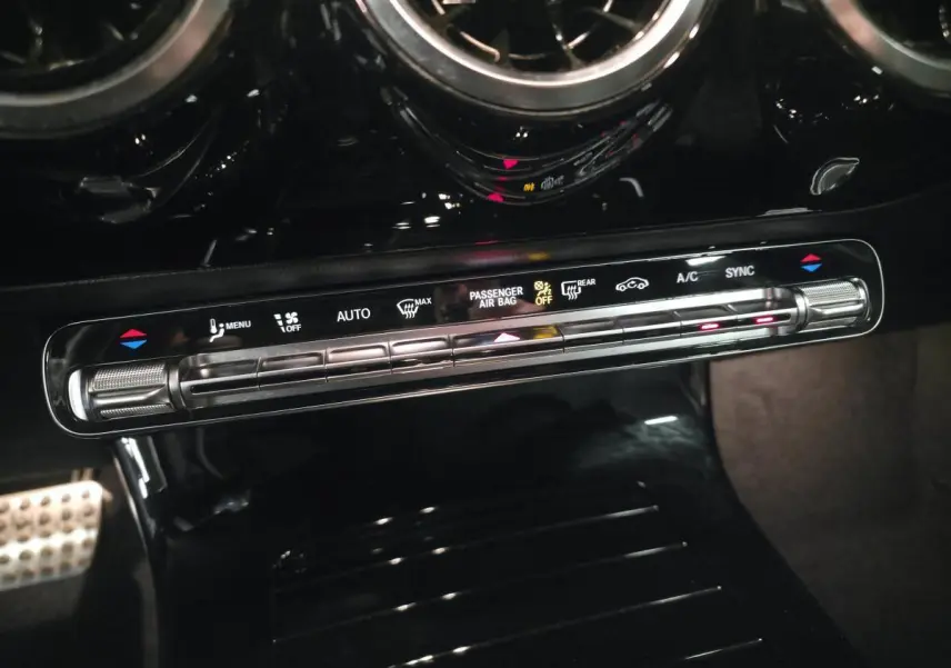 Gros plan sur la console centrale noire brillante avec commandes de climatisation digitale et buses d’aération rondes chromées.