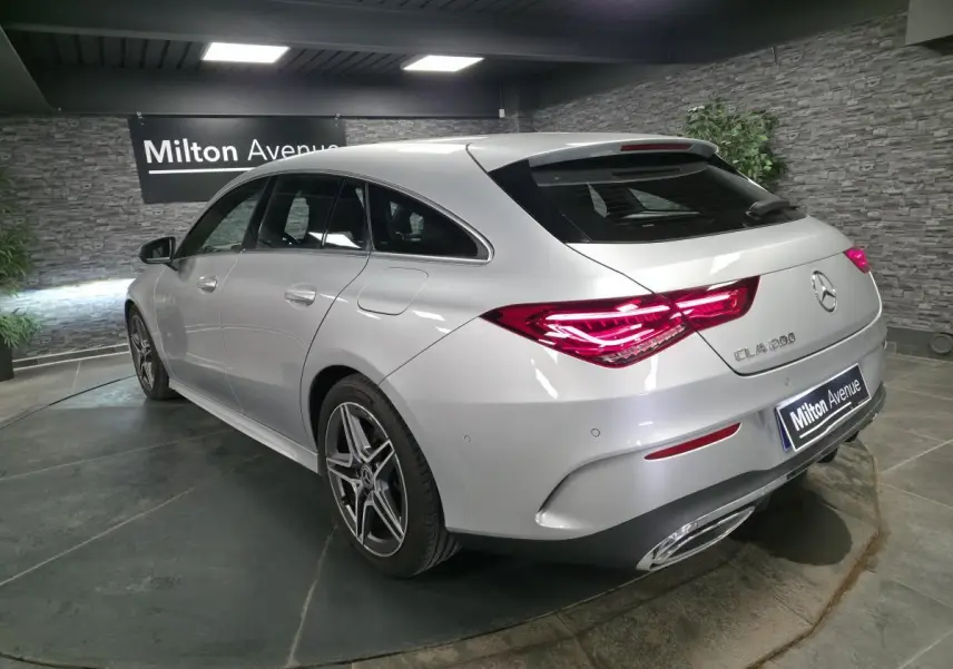 Vue 3/4 arrière droite d'une Mercedes CLA Shooting Brake 200 gris métallisé avec jantes AMG et feux arrière LED allumés.