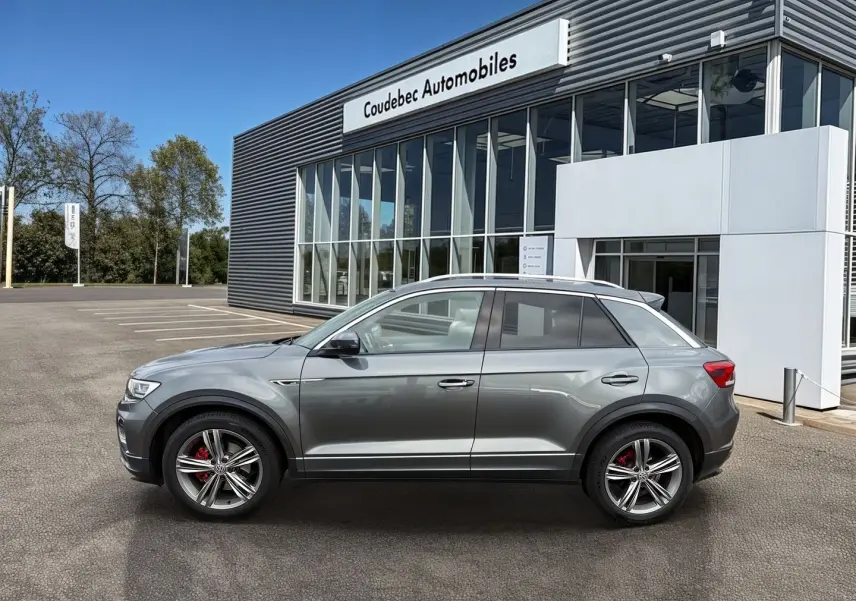 Profil côté gauche d'un Volkswagen T-Roc gris Indium Metal avec jantes alliage et étriers de frein rouges, devant un bâtiment.