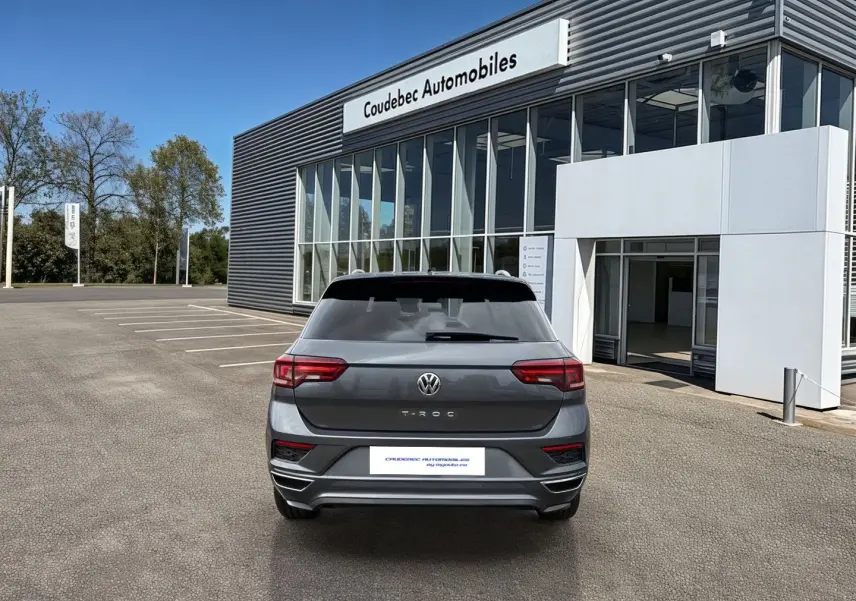 Vue arrière d'un Volkswagen T-Roc gris Indium Metal garé devant un bâtiment moderne de concession automobile.