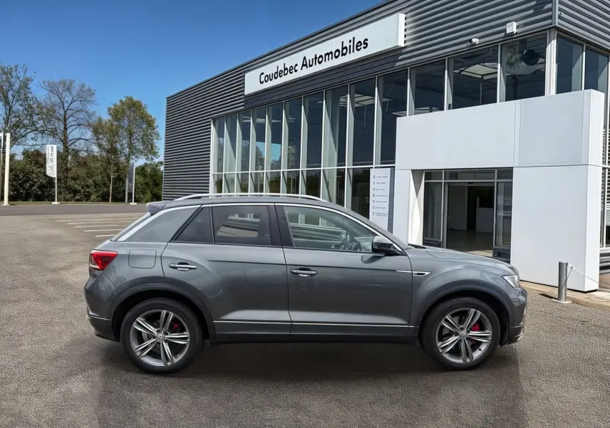 Vue latérale droite d'un Volkswagen T-Roc gris Indium Metal avec jantes alliage et étriers rouges, devant un bâtiment commercial.