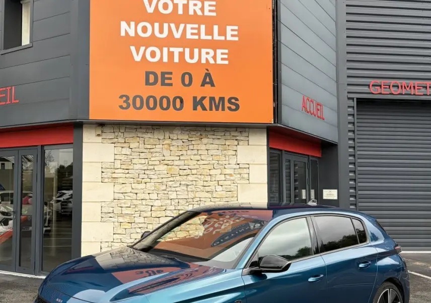 Peugeot 308 bleu 3/4 avant droit, avec feux LED et jantes alliage, devant un bâtiment commercial.