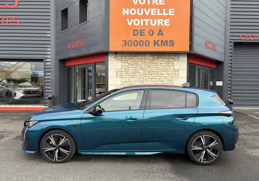 Profil latéral gauche d'une Peugeot 308 bleu métallisé 2025 garée devant un bâtiment avec enseigne orange.