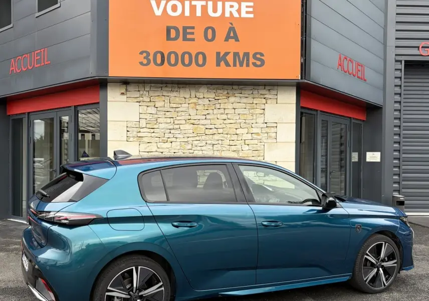 Vue latérale droite d'une Peugeot 308 1.2 Hybrid 145 GT bleu métallisé, garée devant un bâtiment avec enseigne orange.