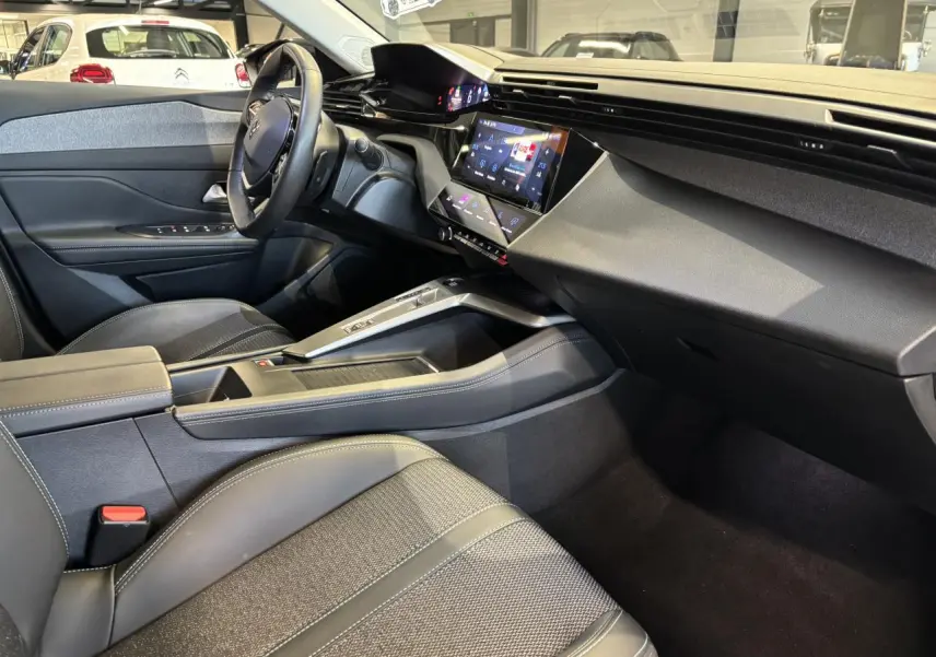Vue intérieure côté conducteur de la Peugeot 308 SW 2024, sellerie tri matières noire avec surpiqûres bleues et tableau de bord moderne.
