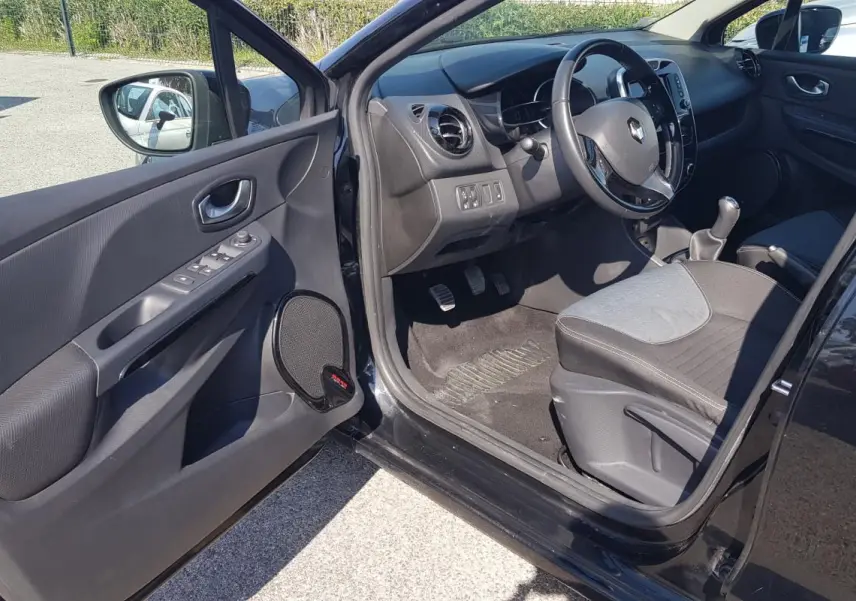 Vue intérieure côté conducteur d'une Renault Clio IV noire, montrant le volant cuir, la boîte manuelle et les commandes de porte.