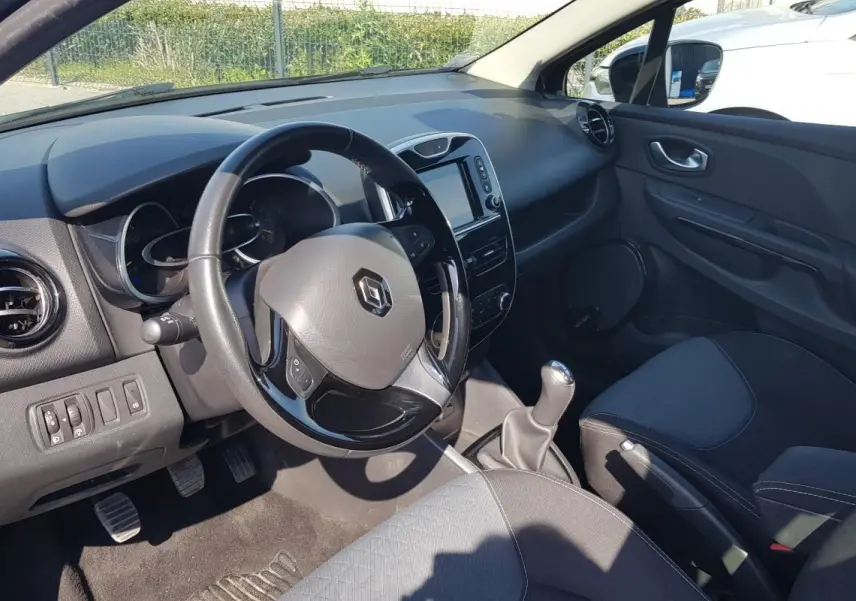 Vue intérieure côté conducteur de la Renault Clio IV noire, montrant le volant cuir, tableau de bord et levier de vitesse manuel.