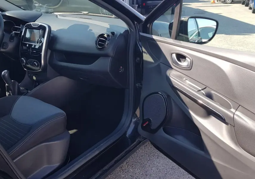 Intérieur avant gauche d’une Renault Clio IV noire, montrant tableau de bord, volant cuir et porte ouverte.