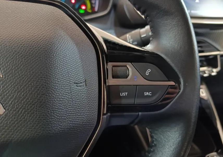 Gros plan sur les commandes droite du volant cuir noir du Peugeot 208 électrique blanc, avec tableau de bord en arrière-plan flou.