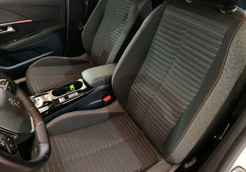 Vue intérieure rapprochée des sièges avant noirs avec surpiqûres rouges et console centrale d’une Peugeot 208 Electrique blanche.