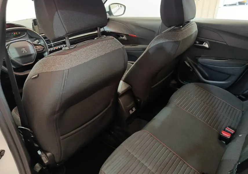 Vue intérieure arrière de la Peugeot 208 électrique blanche, montrant les sièges noirs avec surpiqûres rouges et la console centrale.