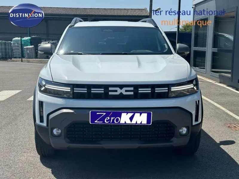 Vue de face du Dacia Duster blanc Glacier 2025 avec calandre noire et logo central distinctif, sur parking extérieur.