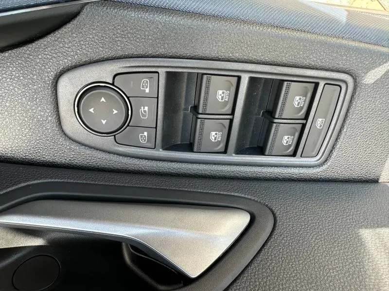 Gros plan sur les commandes des vitres électriques et réglages des rétroviseurs côté gauche intérieur du Dacia Duster blanc 2025.