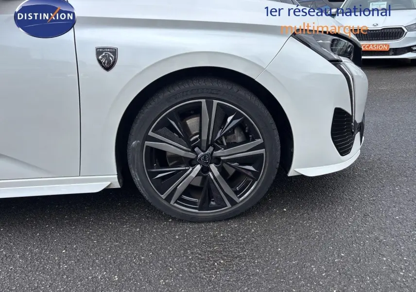 Gros plan sur la roue avant droite et l'aile avant d'une Peugeot 308 SW blanche nacrée, avec logo lion visible.