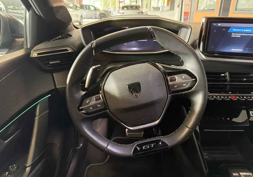 Vue intérieure centrée sur le volant GT de la Peugeot 208 Hybrid 145, avec tableau de bord et écran tactile visibles.