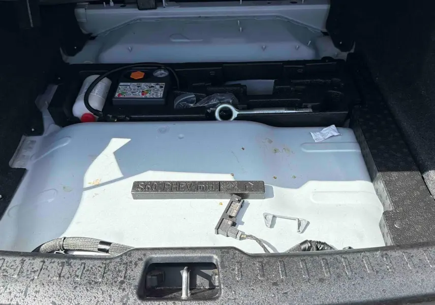 Vue du coffre ouvert d'une Volvo S60 blanche montrant le compartiment de rangement sous plancher avec outils et kit de réparation.