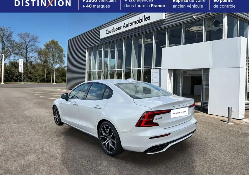 Vue 3/4 arrière droite d'une Volvo S60 blanche avec jantes alliage noir et calandre noire brillante devant un bâtiment commercial.
