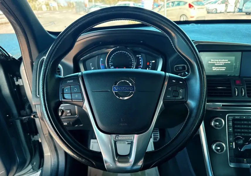 Vue rapprochée du volant noir du Volvo XC60 gris 2016 avec commandes intégrées et tableau de bord digital.