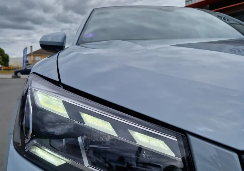 Gros plan sur le phare avant gauche de l'Audi Q2 35 TFSI 2023 gris flèche avec lignes LED distinctives sous ciel nuageux.