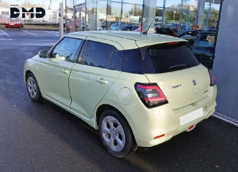 Vue 3/4 arrière droite d'une Suzuki Swift 1.2 Privilège Hybrid jaune clair avec vitres teintées, garée devant un bâtiment vitré.