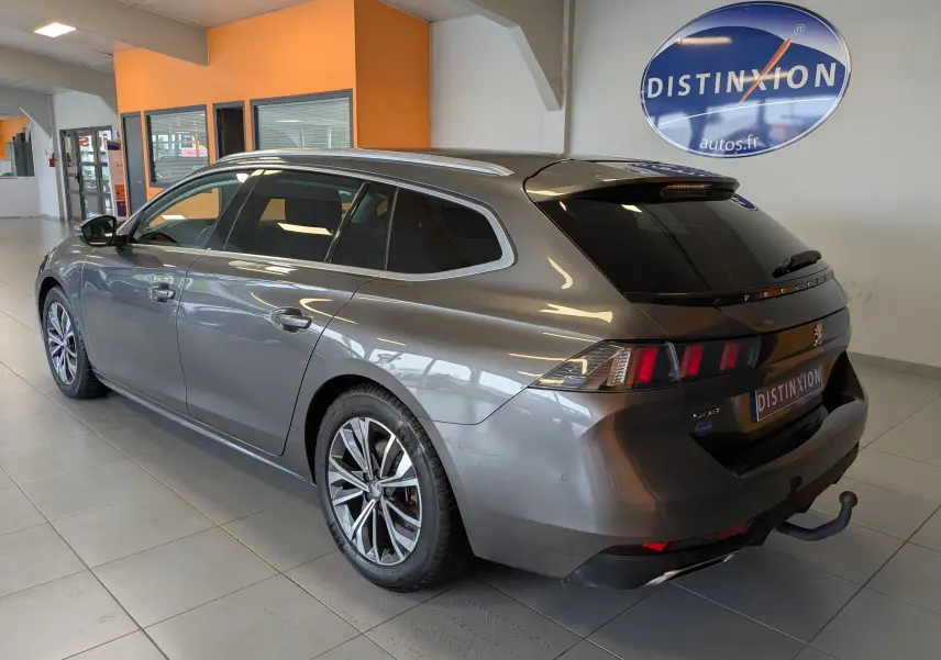 Vue 3/4 arrière droite d'une Peugeot 508 SW gris métallisé avec barres de toit et attelage, stationnée en intérieur showroom.