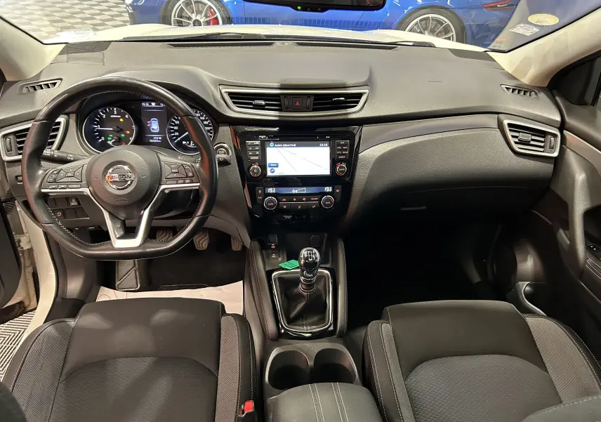 Vue intérieure avant du Nissan Qashqai blanc 2018, montrant le tableau de bord, volant cuir multifonction et levier de vitesse manuel.