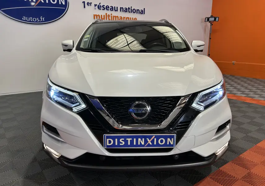 Vue de face d'un Nissan Qashqai blanc avec phares LED allumés et calandre noire brillante dans un showroom.