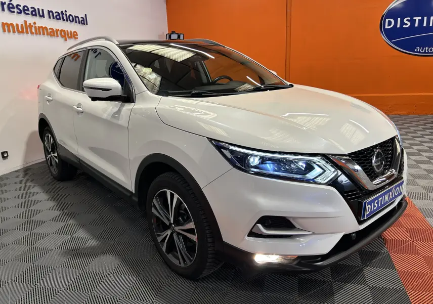 Nissan Qashqai blanc en 3/4 avant droit, avec phares allumés et toit panoramique visible en intérieur showroom.