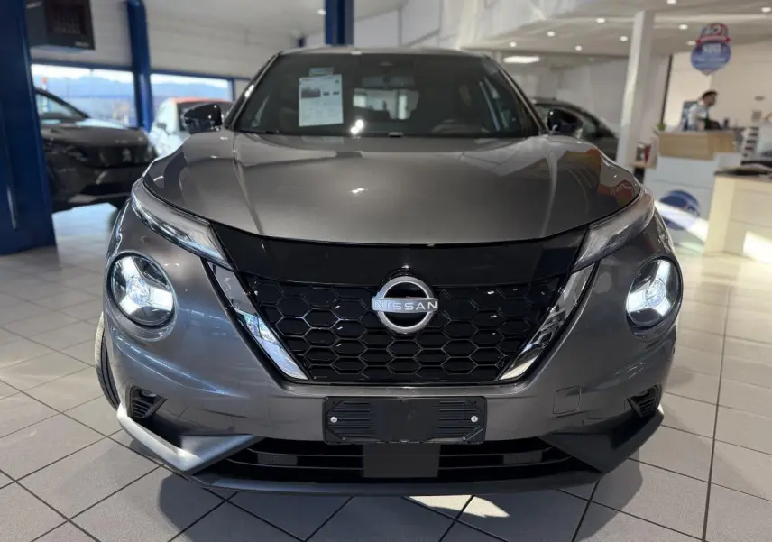 Vue frontale d'un Nissan Juke Hybrid gris métallisé en showroom avec calandre noire et phares LED ronds allumés.