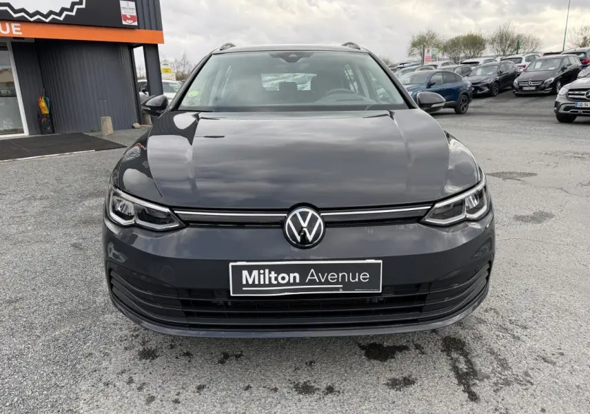 Vue frontale d'une Volkswagen Golf SW gris Urano avec calandre noire et logo VW centré sur un parking extérieur.