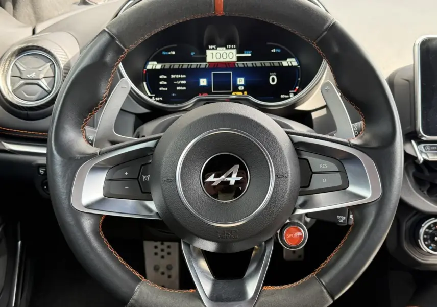 Vue rapprochée du volant noir avec surpiqûres orange et tableau de bord numérique de l'Alpine A110 Gris Tonnerre Mat.