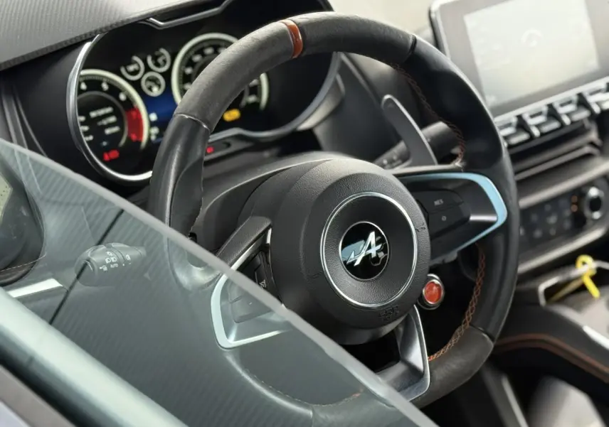 Gros plan sur le volant cuir noir avec surpiqûres orange de l'Alpine A110 1.8T 292 S, tableau de bord flou en arrière-plan.