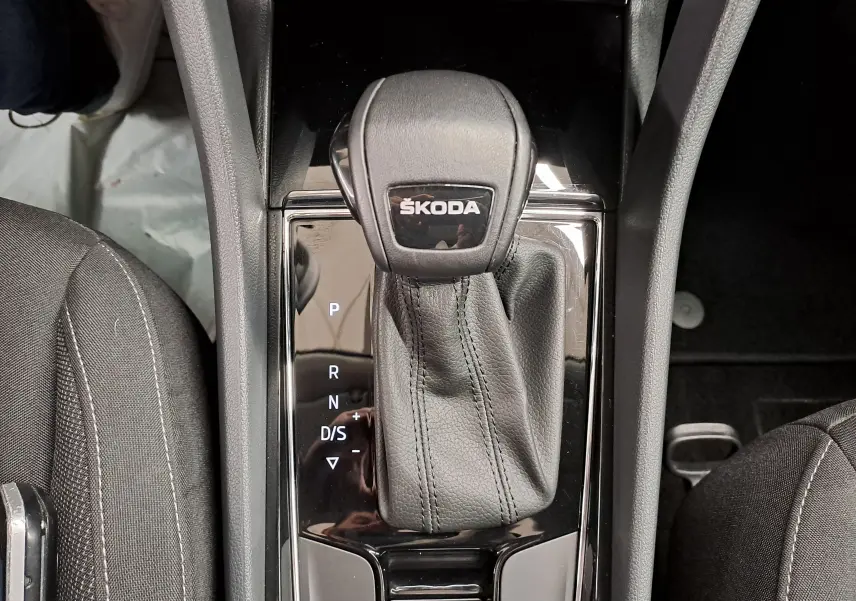 Gros plan sur le levier de vitesses automatique noir avec logo ŠKODA, entouré des sièges avant en tissu gris foncé.