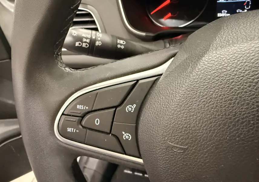 Gros plan sur les commandes de régulateur de vitesse au volant noir de la Renault Mégane IV 1.5 dCi 115 gris Highland, tableau de bord en arrière-plan.