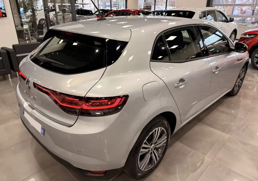 Vue 3/4 arrière droite d'une Renault Mégane IV gris Highland dans un showroom avec d'autres véhicules visibles.