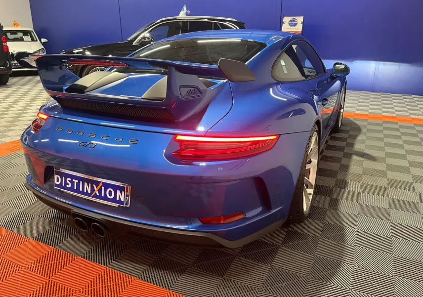 Vue 3/4 arrière droite d'une Porsche 911 GT3 bleu saphir avec grand aileron et feux arrière allumés.