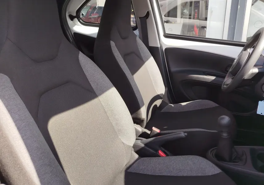 Vue intérieure côté conducteur de la Toyota Aygo X blanche, montrant les sièges tissu bicolore et la boîte manuelle.