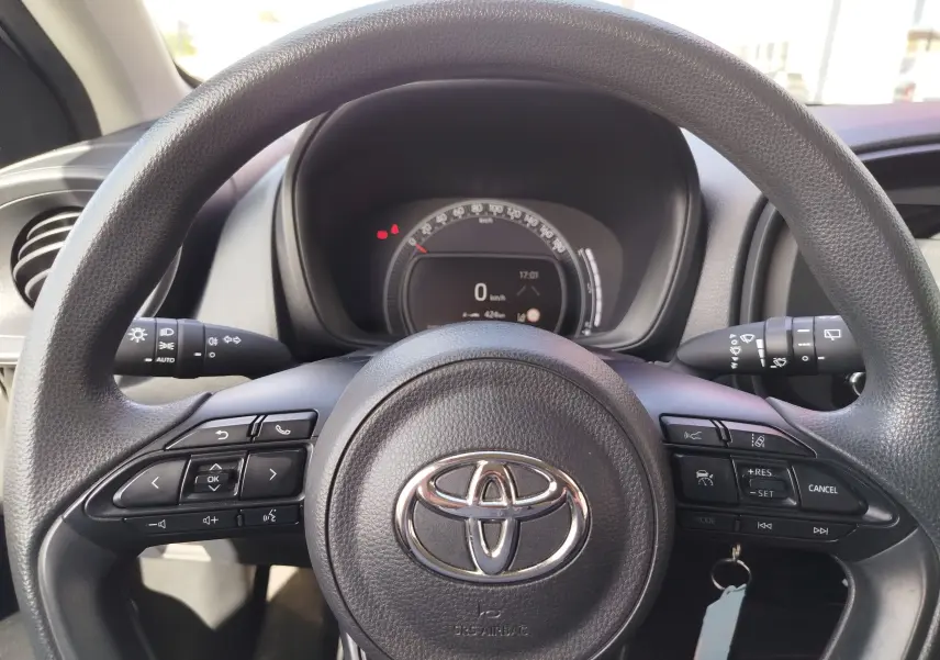 Vue rapprochée du volant noir de la Toyota Aygo X 2023, avec commandes multifonctions et tableau de bord digital en arrière-plan.