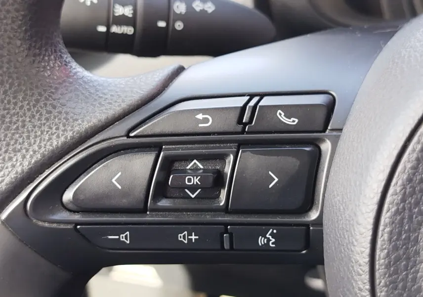Gros plan sur les commandes noires du volant côté gauche de la Toyota Aygo X blanche, avec boutons de navigation et appel.
