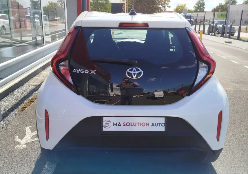 Vue arrière d'une Toyota Aygo X blanche avec hayon noir brillant et feux arrière en forme de boomerang.