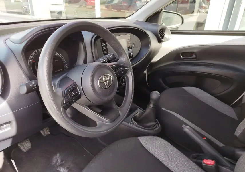 Vue intérieure côté conducteur de la Toyota Aygo X 2023, volant multifonction, sièges tissu noir et gris, levier de vitesse manuel.