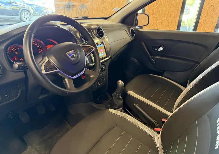 Vue intérieure côté conducteur du Dacia Sandero Stepway 2019, montrant le volant, tableau de bord et sièges noirs avec surpiqûres grises.
