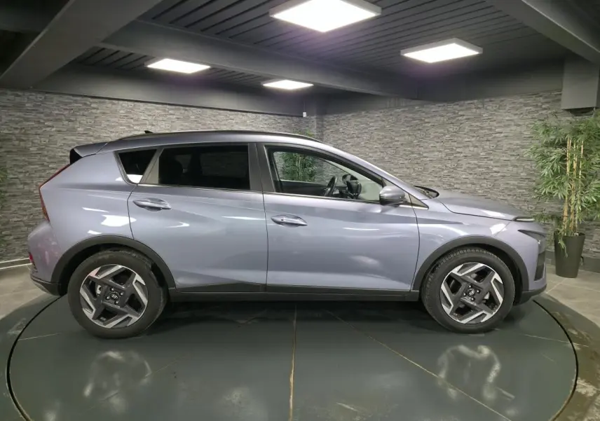 Vue de profil côté gauche d'une Hyundai Bayon gris métallisé 2025 avec jantes alliage et vitres surteintées dans un showroom.